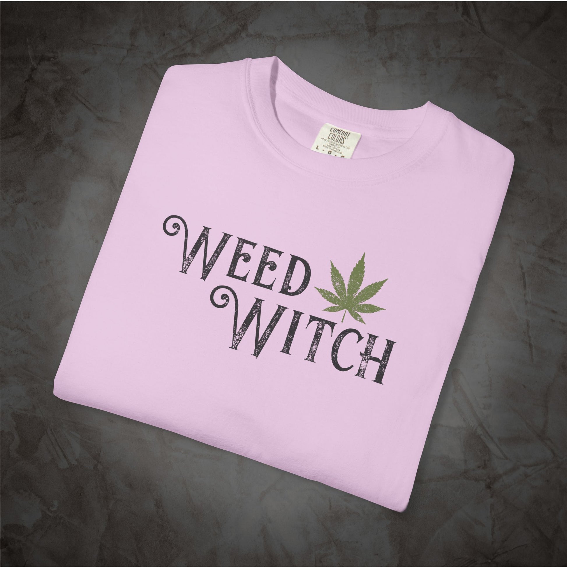 A light pink folded t-shirt with a 'WEED WITCH' graphic printed in black with a green cannabis leaf, displayed on a dark textured background.