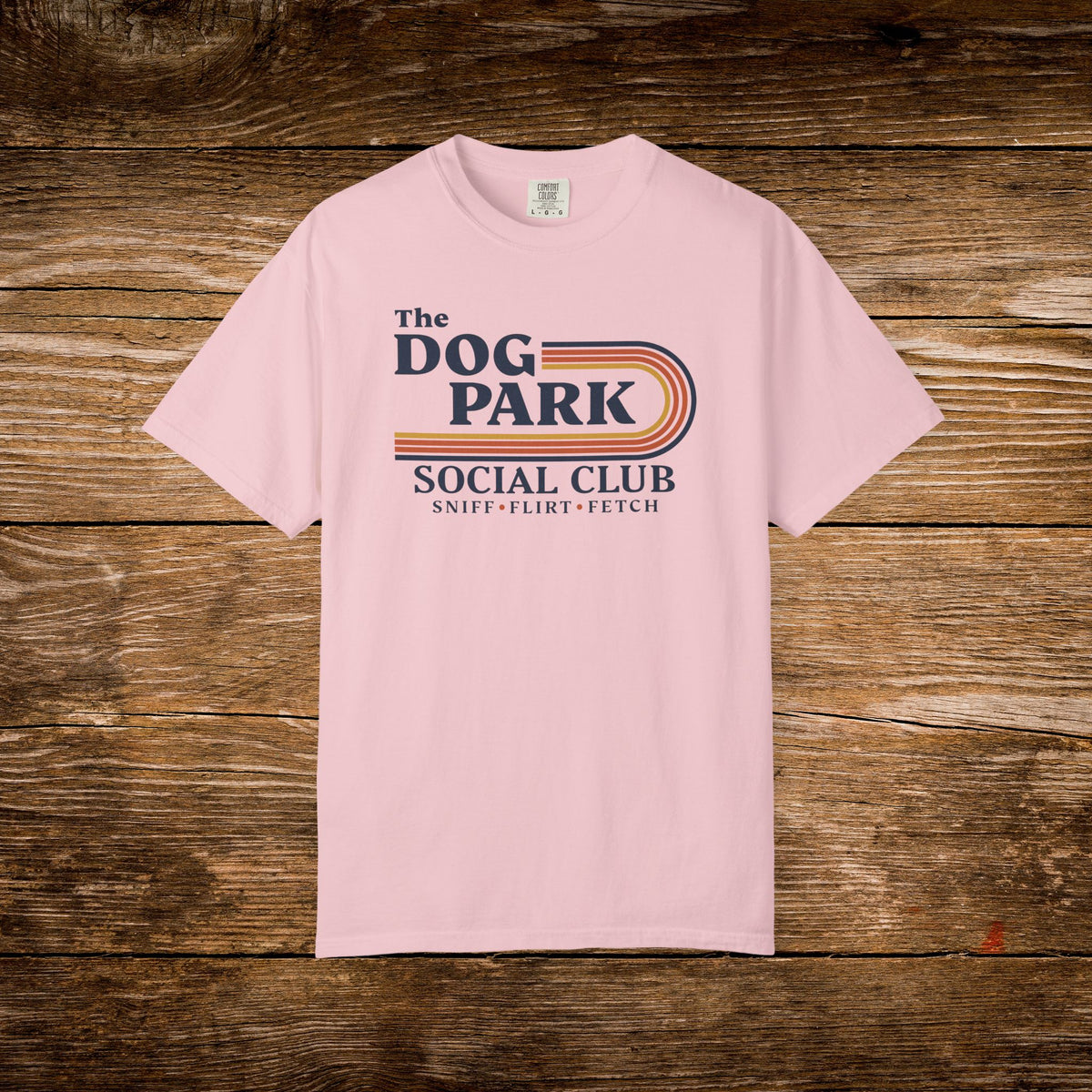 A light pink short-sleeve t-shirt with a retro-style graphic reading 'The DOG PARK SOCIAL CLUB' and 'SNIFF - FLIRT - FETCH' in navy and orange, laid flat on a rustic brown wooden plank background.