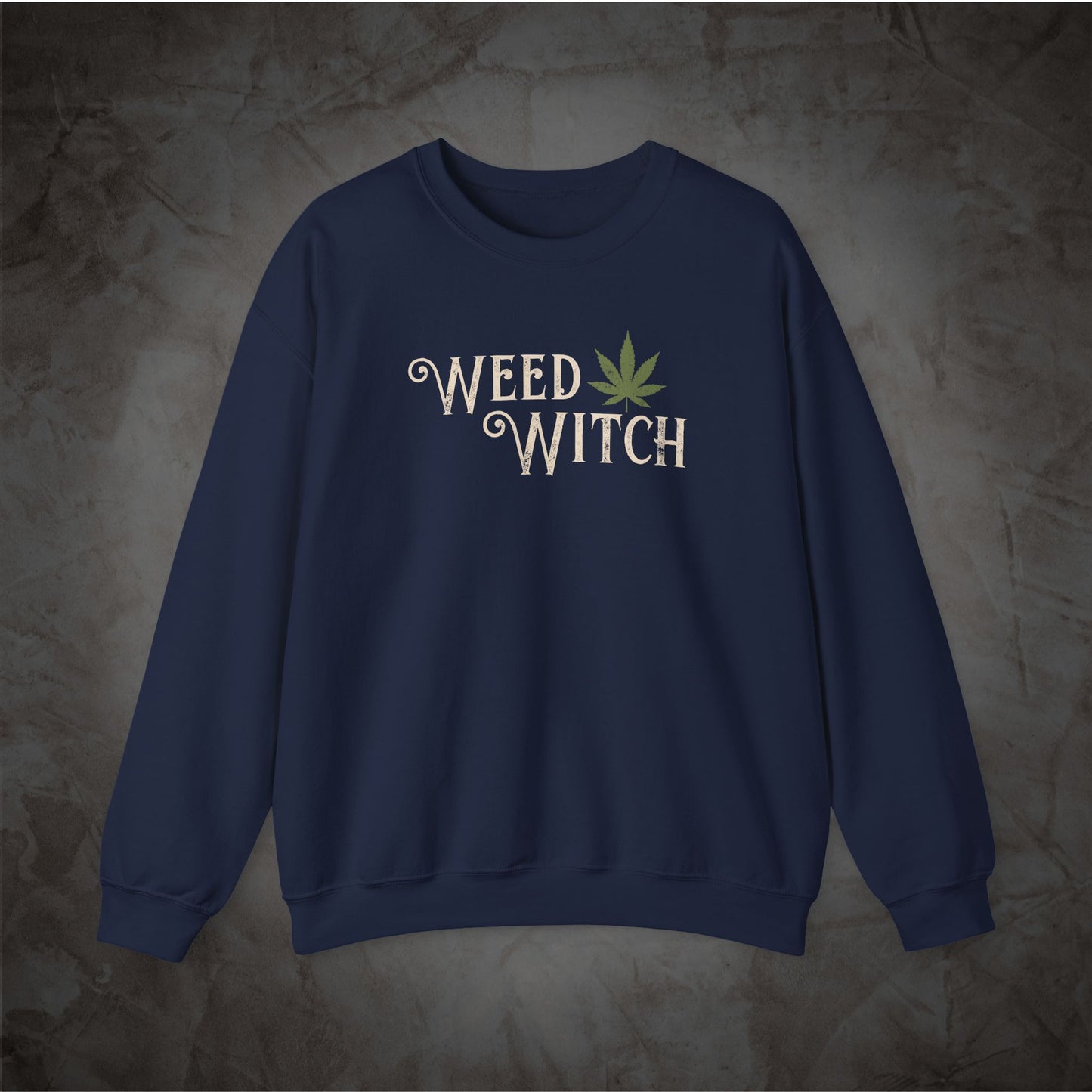 A navy blue crewneck sweatshirt featuring a graphic on the chest that reads 'WEED WITCH' in a white decorative font, with a green cannabis leaf integrated into the design, displayed against a textured gray background.