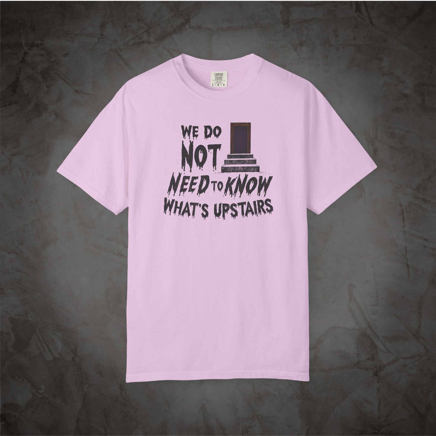 A light pink short-sleeve t-shirt with a black graphic text print reading 'WE DO NOT NEED TO KNOW WHAT'S UPSTAIRS' alongside a top hat illustration, displayed against a dark textured gray background.