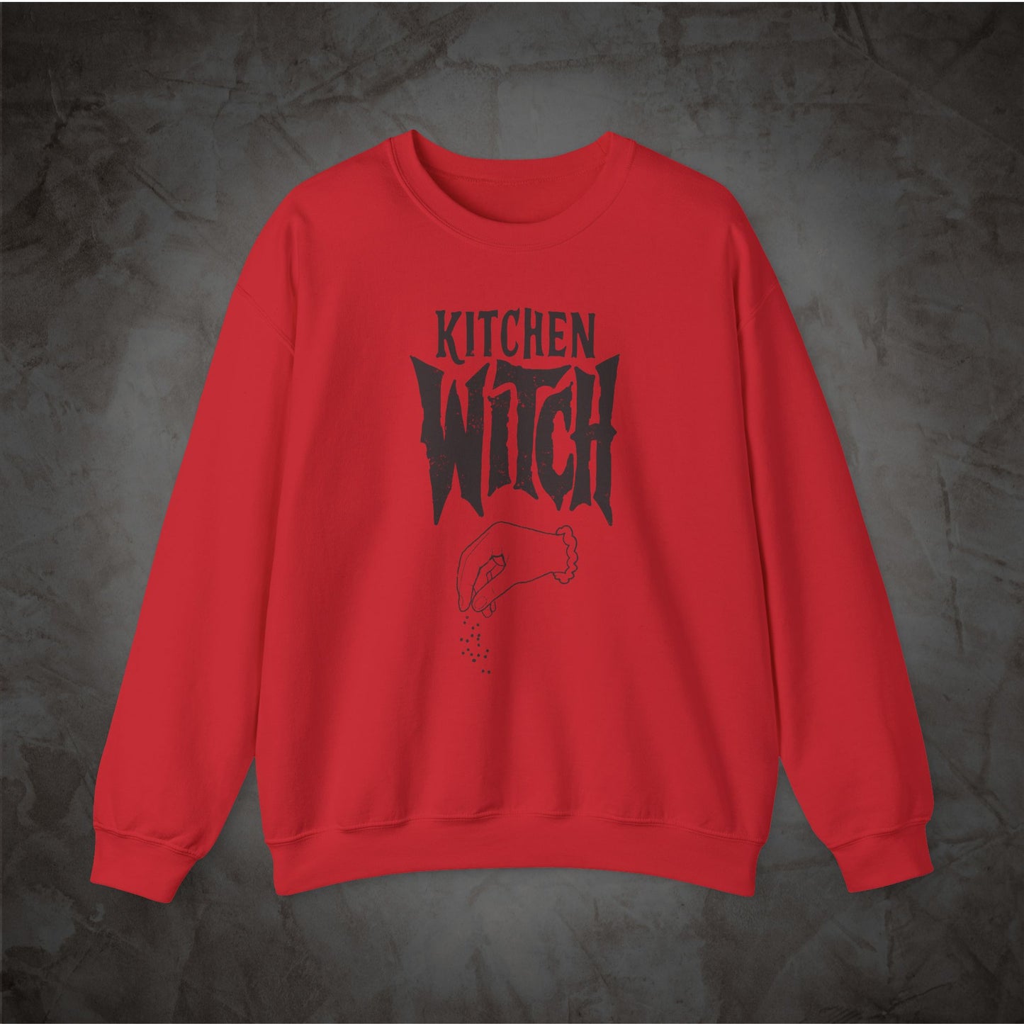 A red crewneck sweatshirt with a black graphic print reading 'KITCHEN WITCH' and a small witch hat illustration, displayed against a dark textured gray background.