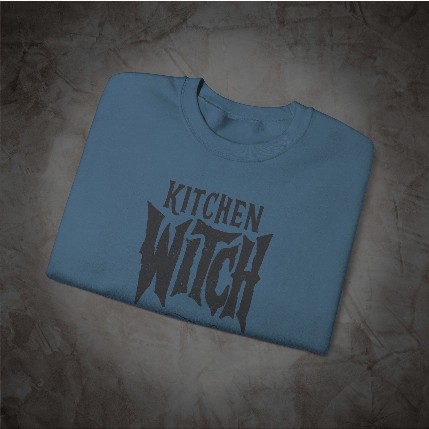 A folded light blue crewneck t-shirt with a dark gray graphic reading 'KITCHEN WITCH' in a stylized font, displayed on a textured brown background.