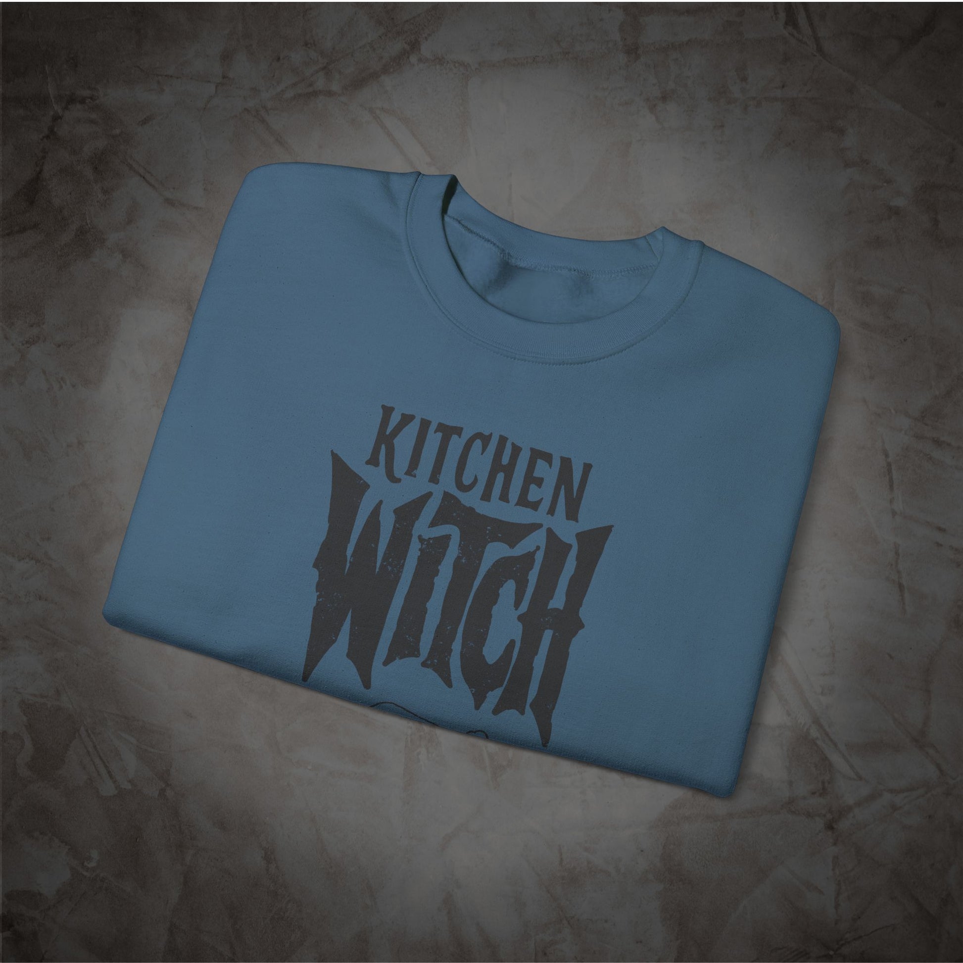 A folded light blue crewneck t-shirt with a dark gray graphic reading 'KITCHEN WITCH' in a stylized font, displayed on a textured brown background.