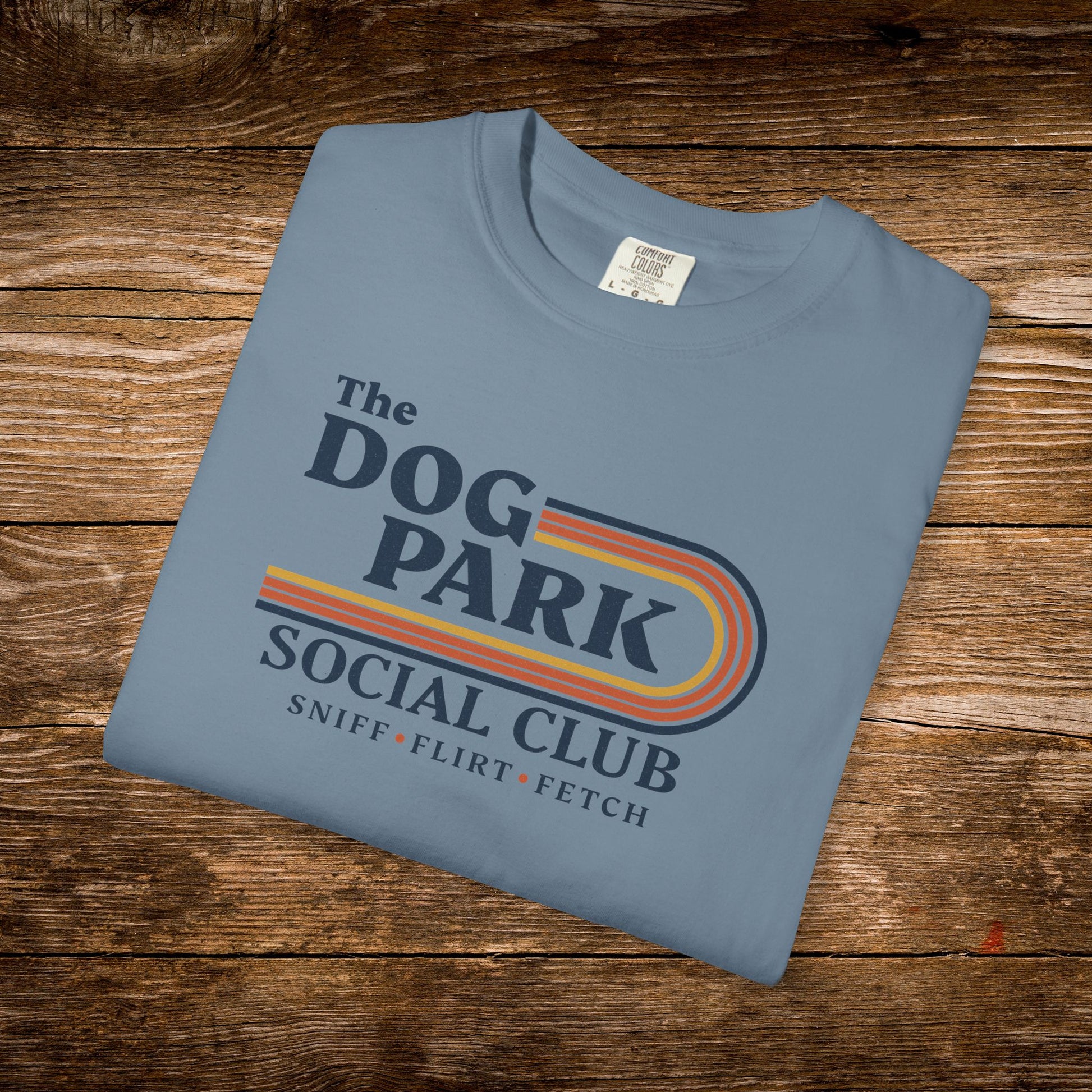 A folded light blue t-shirt with a retro-style graphic reading 'The DOG PARK SOCIAL CLUB' and 'SNIFF * FLIRT * FETCH' in dark blue text with orange and yellow stripes, displayed on a rustic wooden background. The design text reads "Sniff, Flirt, Fetch."