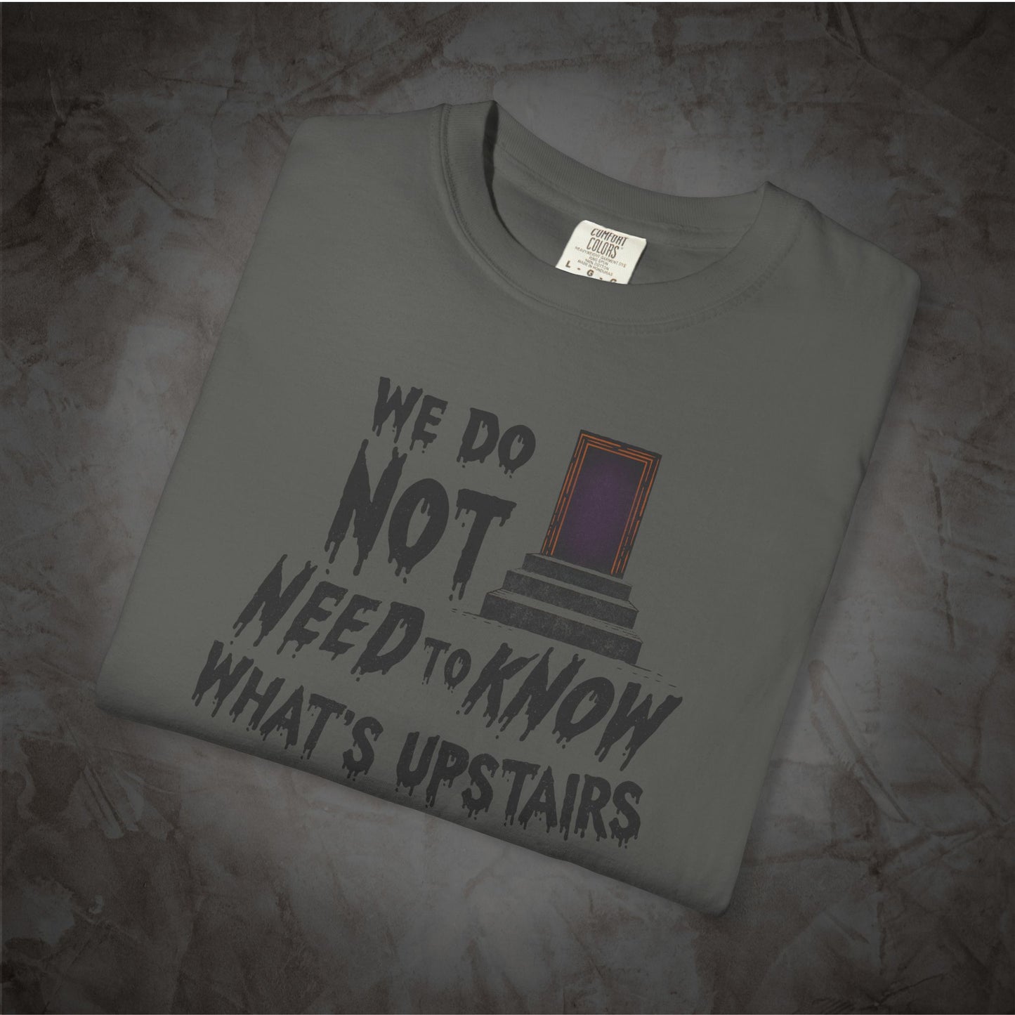 A folded gray t-shirt with a graphic print that reads 'WE DO NOT NEED TO KNOW WHAT'S UPSTAIRS' in a distressed black font, next to a purple and orange door illustration. The shirt is displayed on a textured, dark gray stone-like surface.