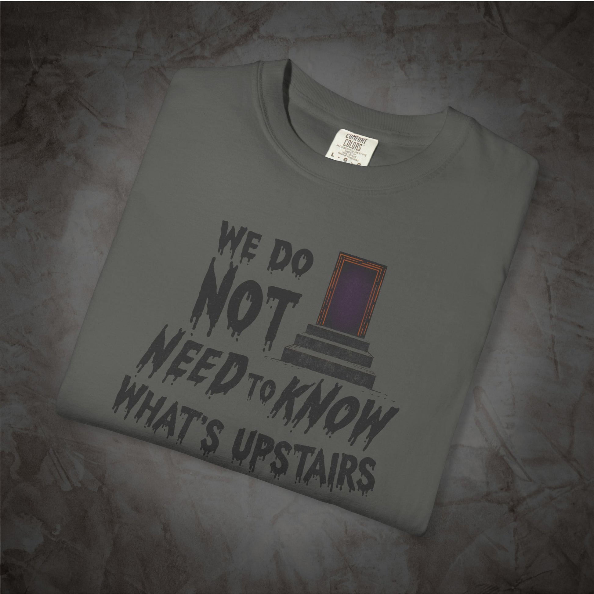 A folded gray t-shirt with a graphic print that reads 'WE DO NOT NEED TO KNOW WHAT'S UPSTAIRS' in a distressed black font, next to a purple and orange door illustration. The shirt is displayed on a textured, dark gray stone-like surface.