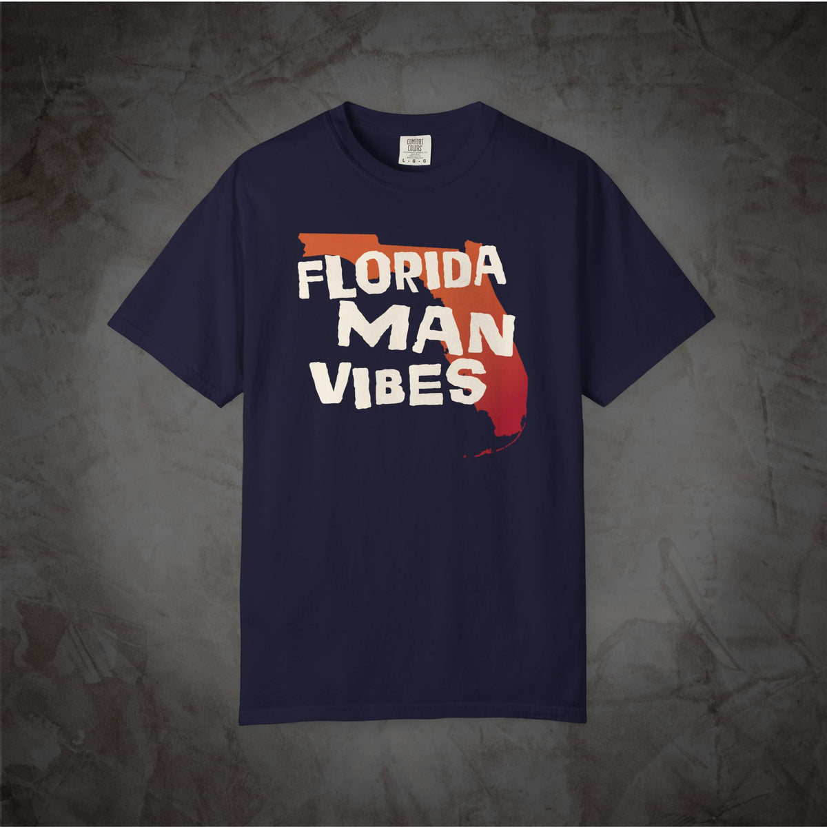 A navy blue short-sleeve t-shirt with a graphic on the chest featuring the text 'FLORIDA MAN VIBES' in white over a red and orange Florida state outline, displayed against a textured gray background.
